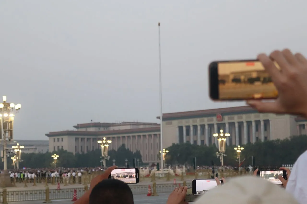 A crowd holding up phones at dawn in Tiananmen Square, in front of a tall flagpole